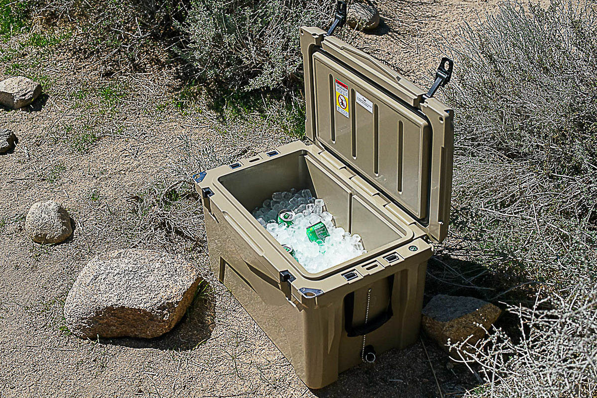 The XSpec 60QT cooler sits on the ground with its lid open, revealing a layer of ice cubes along with several canned beverages.