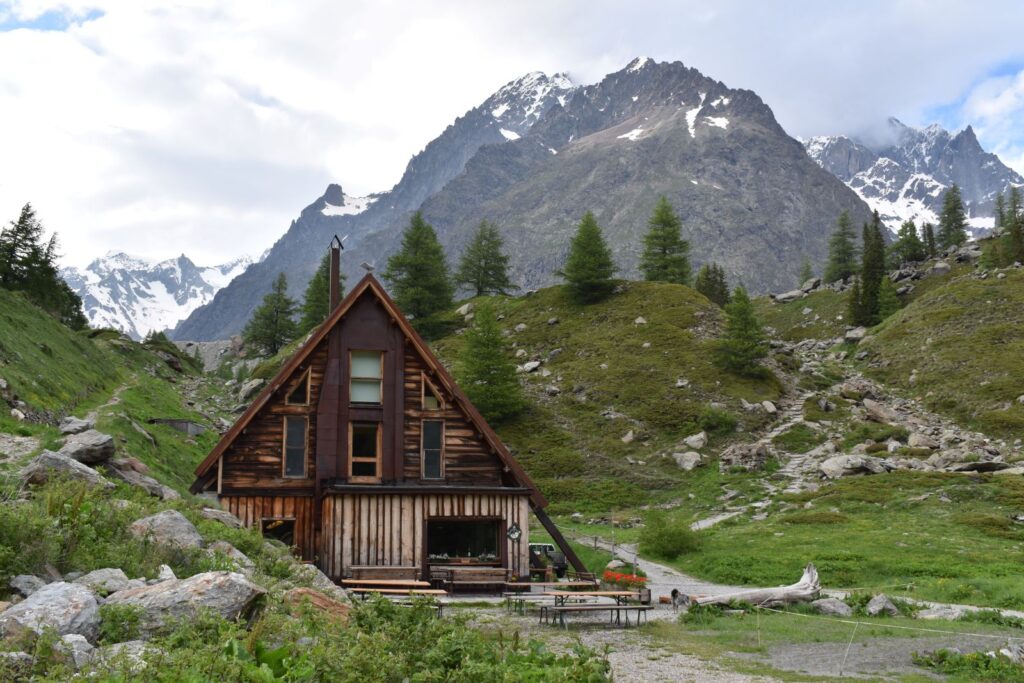 An A-frame cabin is tucked into a green hillside with larger mountains behind it