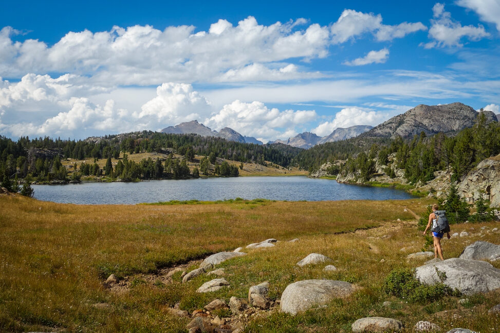 a backpacker hikes through a meadow next to a lake in the wind river range on a partly sunny day