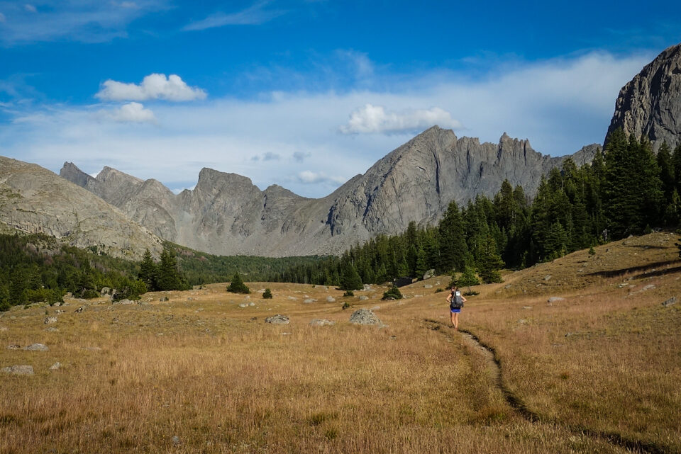 a trail goes through a meadow that a backpacking walks on heading toward granite towers of the wind river range