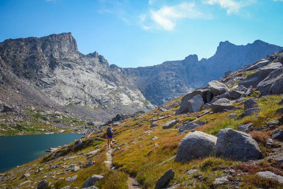 a trail cut in on an alpine meadow hillside with a glittery lake below and granite towers in the background