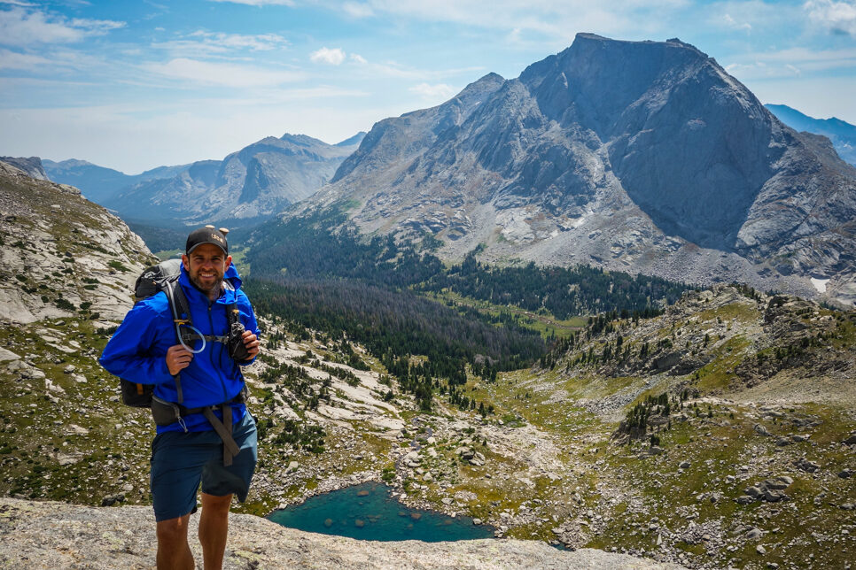 a backpacker posing in front of a clear turquoise lake and big granite mountain on the cirque of the towers backpacking loop