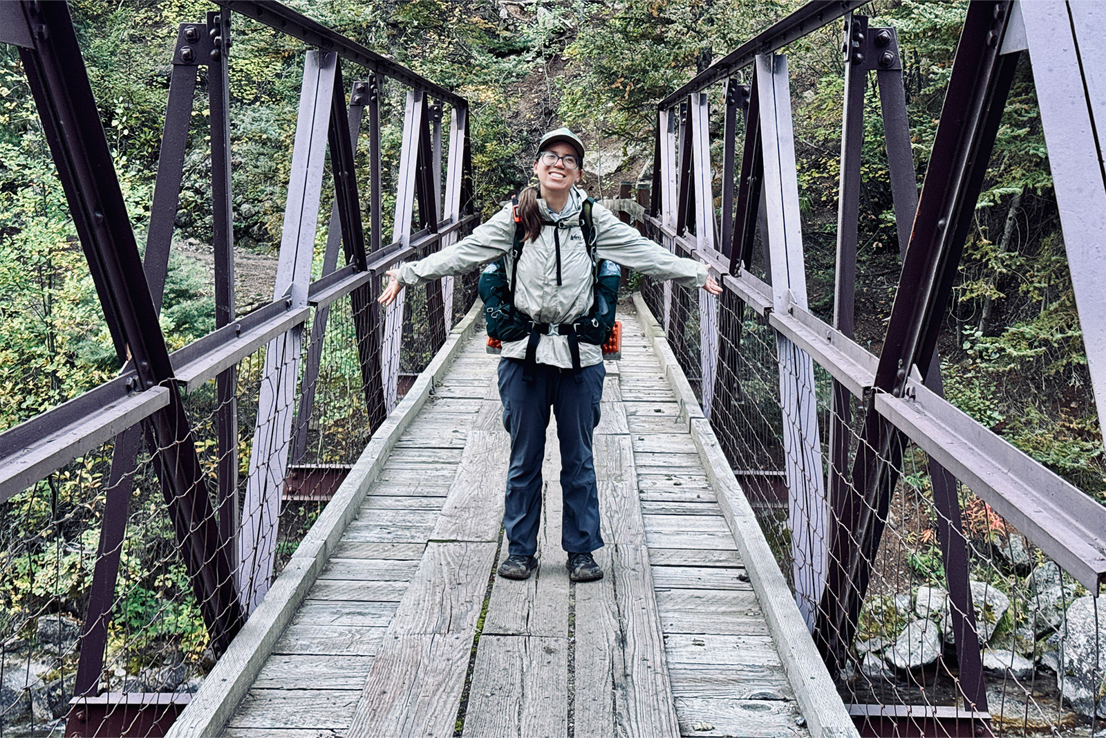 A person wearing the Duluth pants and a backpack is standing on a bridge in the woods looking at the camera.