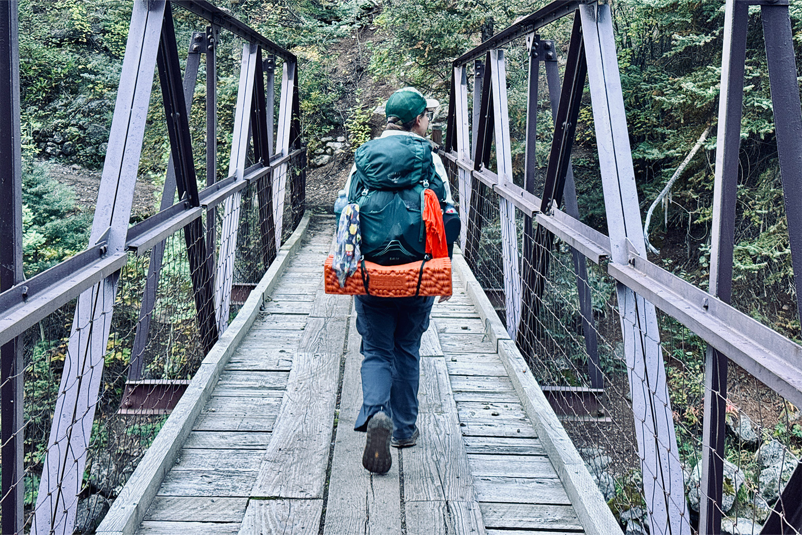 A person wearing the Duluth pants and a backpack is walking across a bridge in the woods.