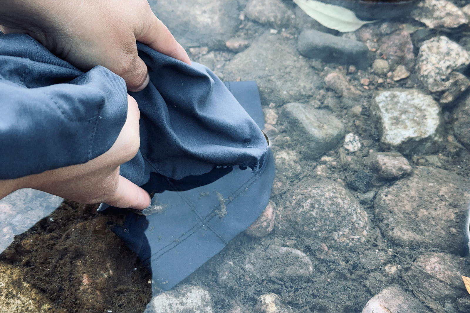 A close up of two hands holding the end of the Duluth pant leg under water in a stream.