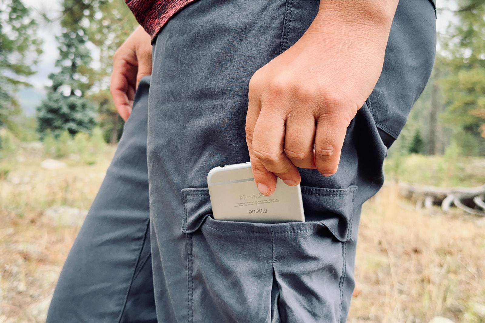 Close up of a persons hand placing a phone in the leg pocket of the Duluth pants. There are pine trees in the background.