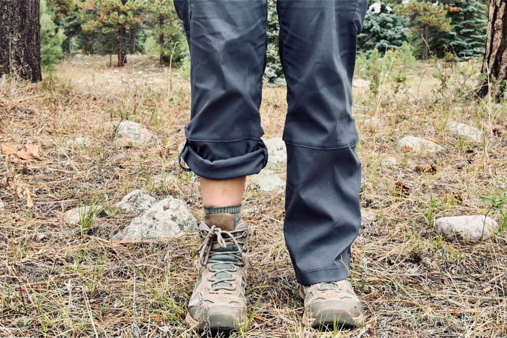 View of a person's lower legs. The leg on the left features the Duluth pants rolled up with the snap feature. The leg on the right demonstrates the pants when worn rolled down. They are wearing boots and standing in a pine forest.