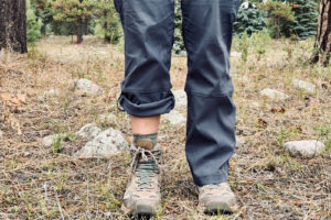 View of a person's lower legs. The leg on the left features the Duluth pants rolled up with the snap feature. The leg on the right demonstrates the pants when worn rolled down. They are wearing boots and standing in a pine forest.