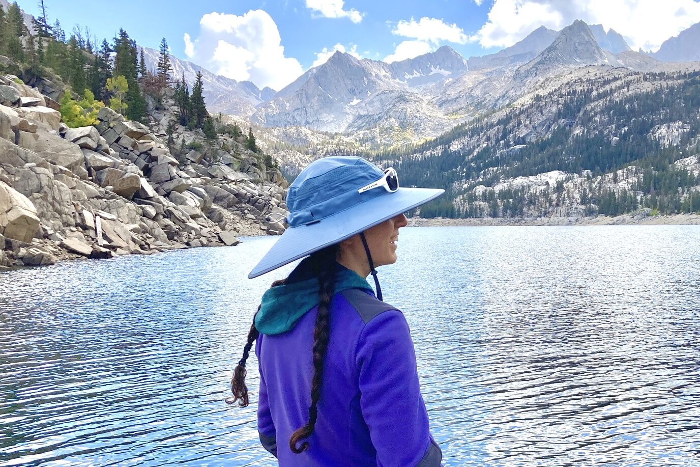 A woman looks out at mountain peaks in the distance in front of a lake, wearing her sun hat.
