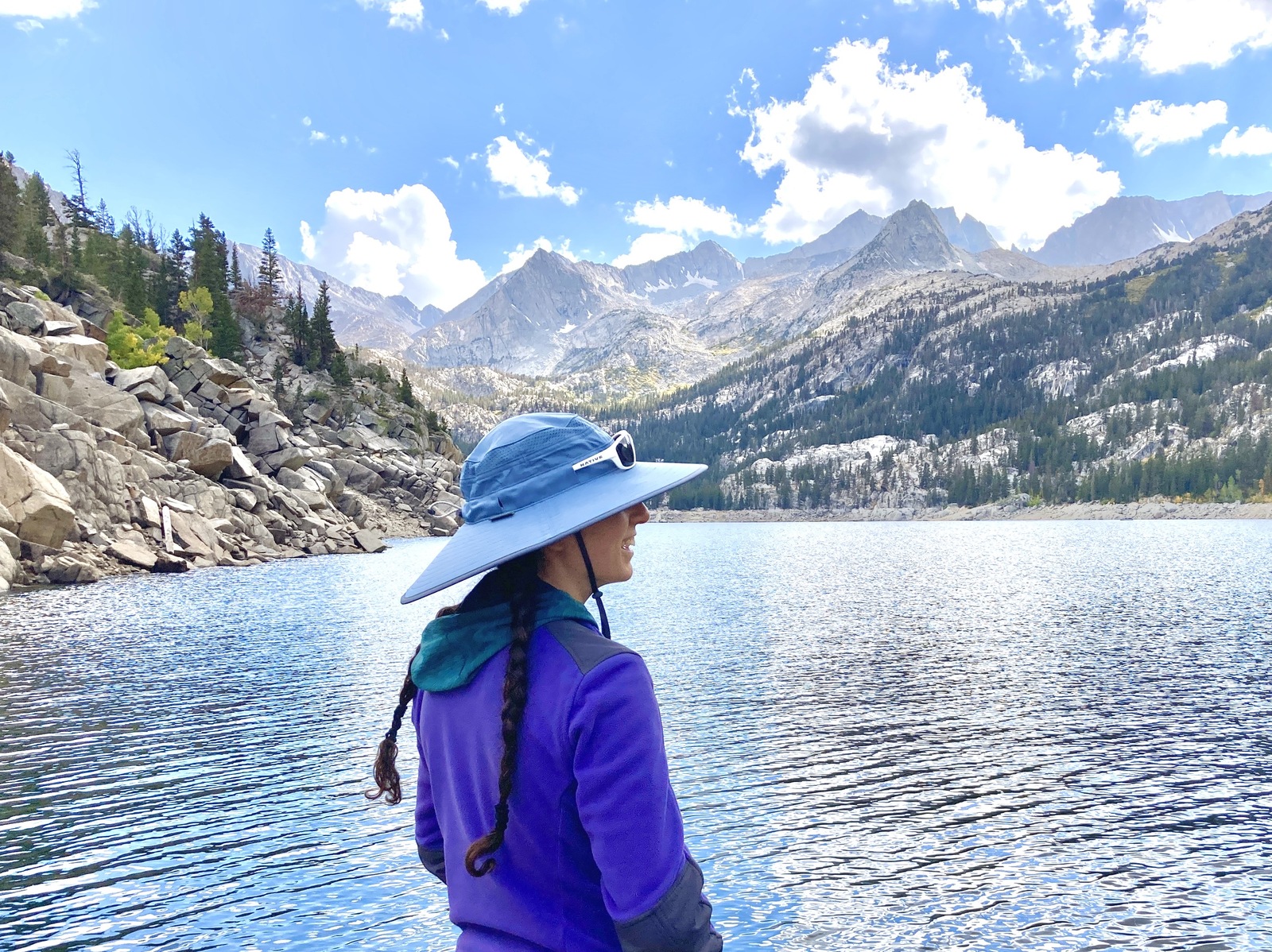 A woman looks out at mountain peaks in the distance in front of a lake, wearing her sun hat.