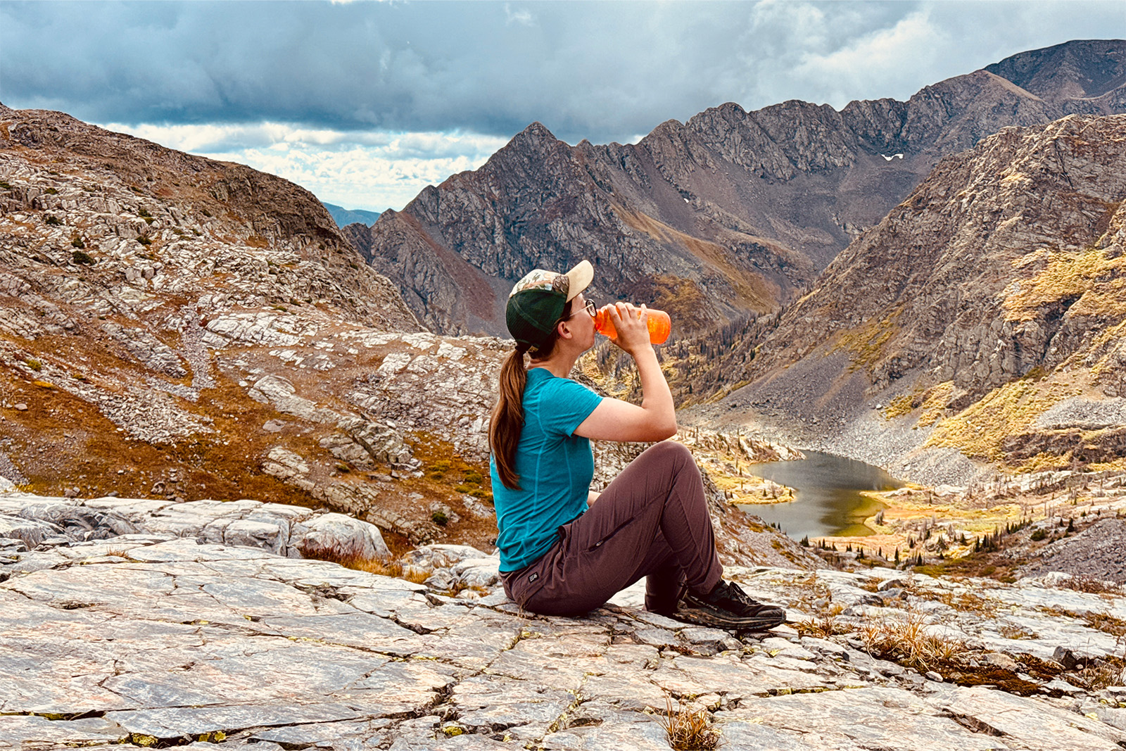 A person wearing the Dynama pants is sitting on a rock on a mountain ridge drinking some water.