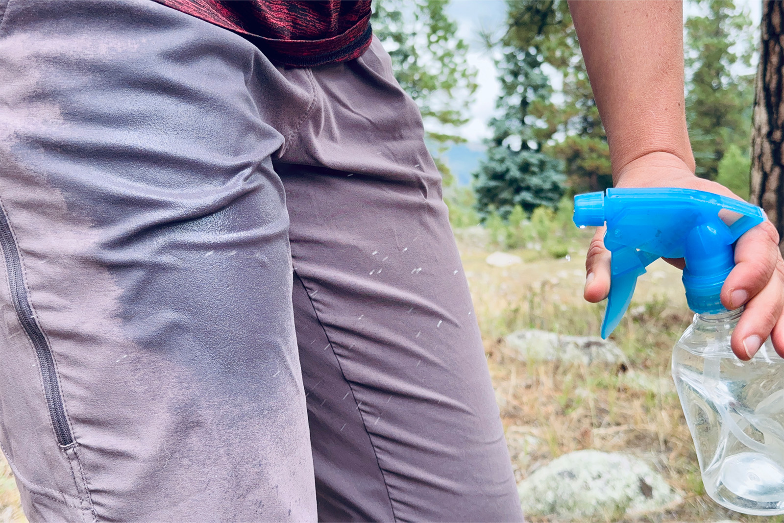 A close up shot of a hand spraying water on the thigh area of the Dynama pants with a squirt bottle. There are some pine trees in the background.