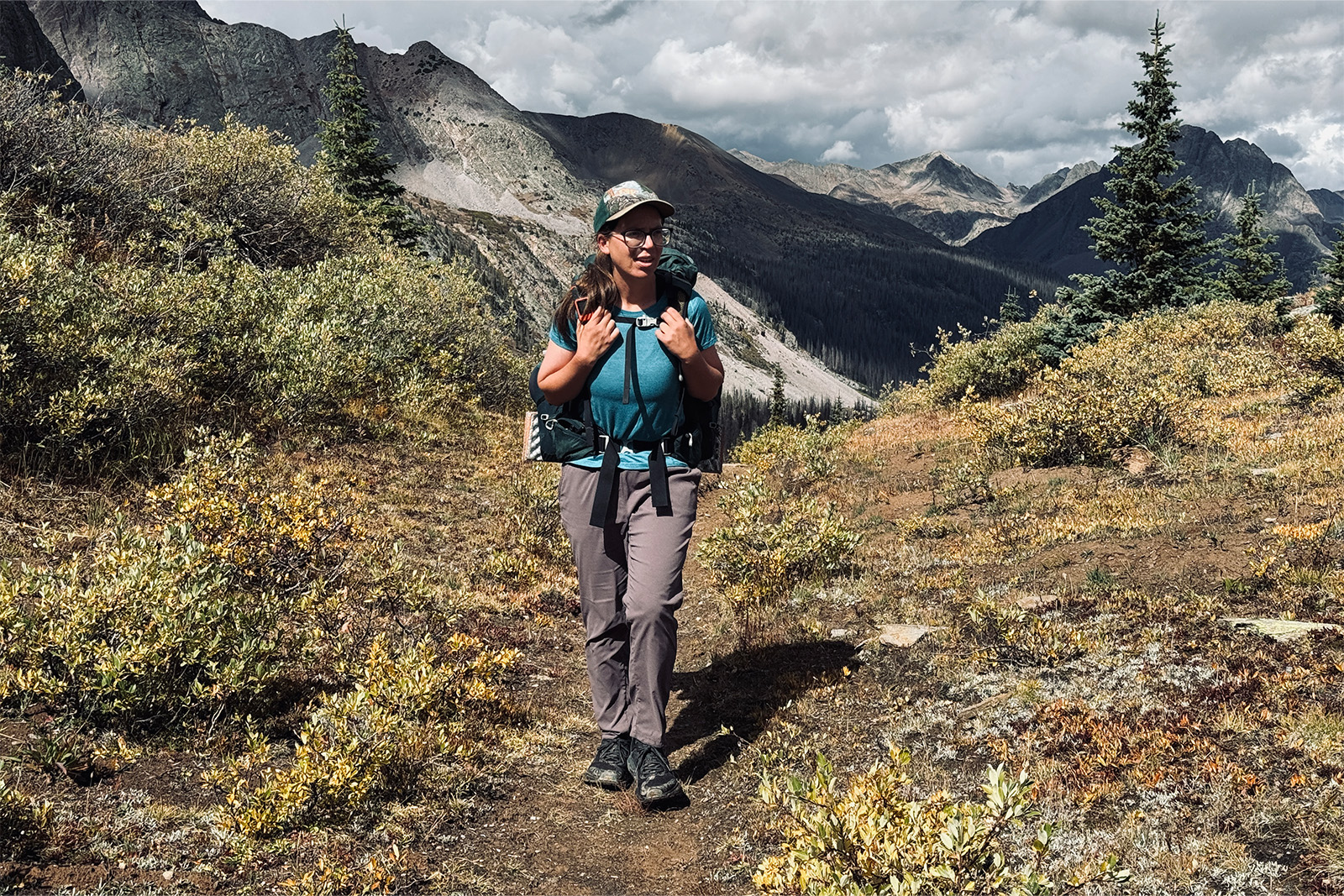 A person wearing the Dynama pants is wearing a backpacking and walking on a trail in the mountains.