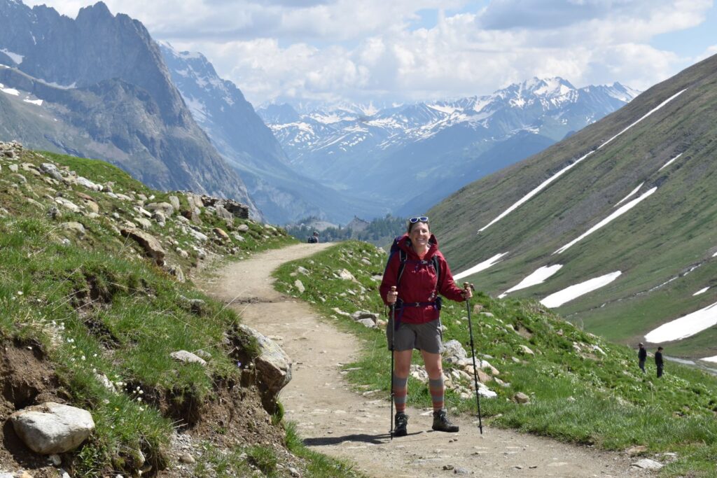 A female hiker in a red jacket poses on an easy trail that descends into an Italian valley surrounded by snowcapped mountains