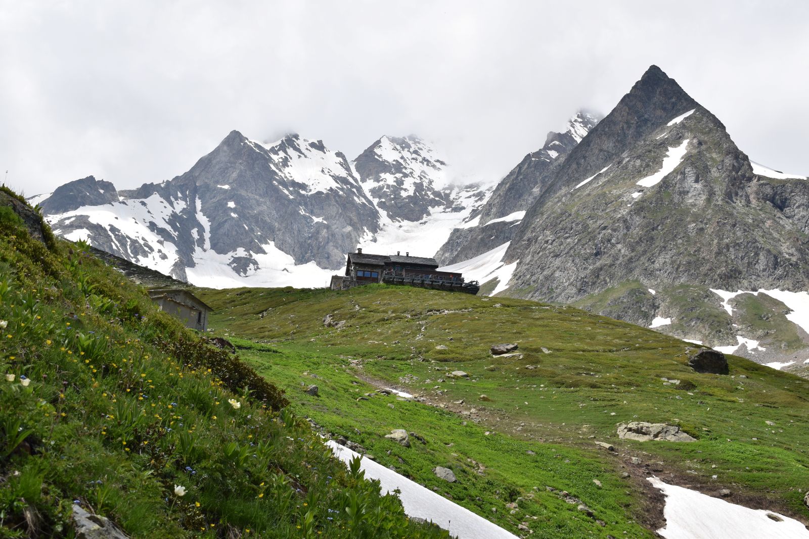 A mountain refuge sits on a green hillside flanked by snowy mountains
