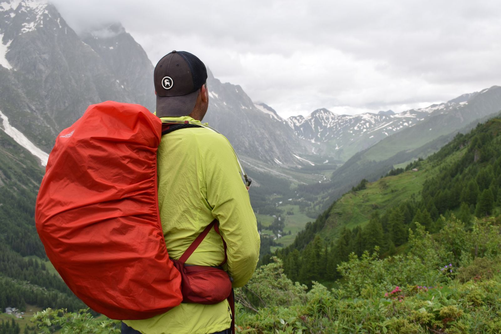 A hiker looks down into a misty mountain valley while wearing backwards ball cap and day pack with a rain covered