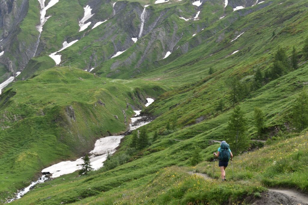 A hiker descends the TMB through lush green meadows streaked with spring snow that hasn't quite melted