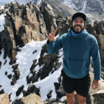 A smiling man flashes a peace sign on top of a snowy mountain.