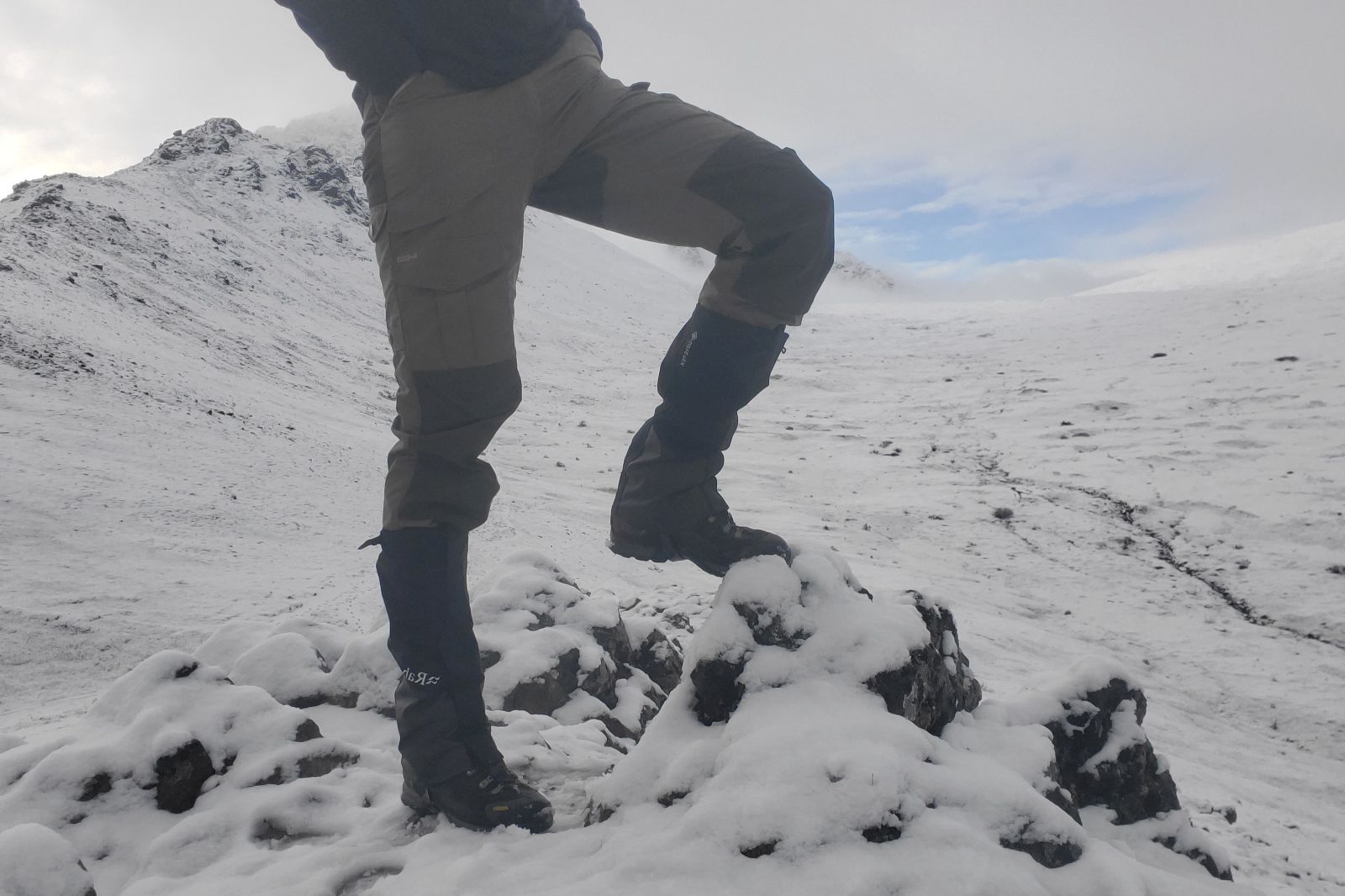 A man stands on a snowy mountain top showing hiking gaiters.