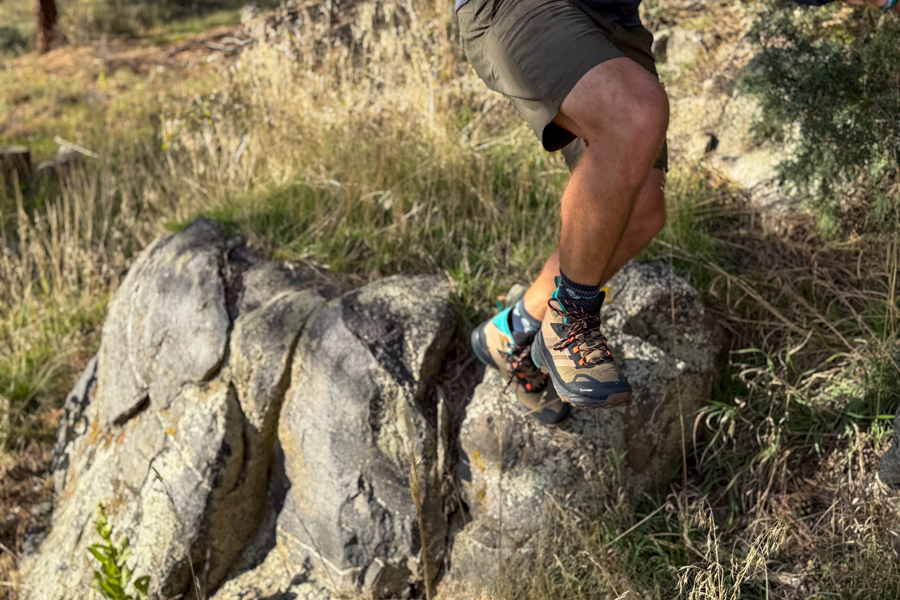 Hiker wearing Adidas Terrex Skychaser AX5 Mid GORE-TEX Climawarm+ boots jumping between two large rocks in a dry grassy landscape.