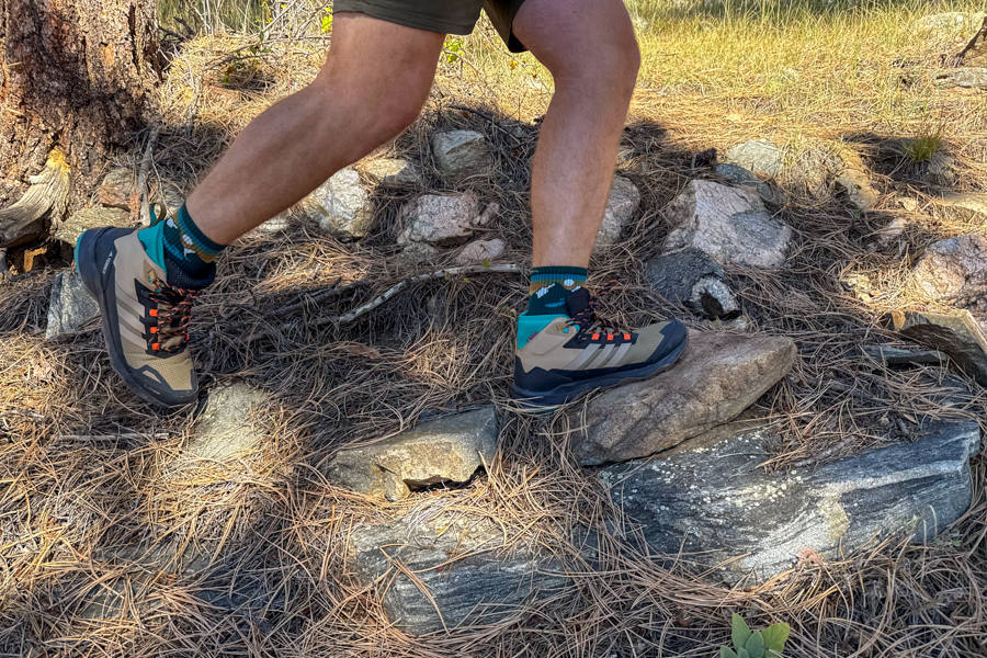 A hiker steps confidently across a rocky trail wearing Adidas Terrex Skychaser AX5 Mid GORE-TEX Climawarm+ hiking shoes. The shoes feature a tan and black design with teal and orange accents, and the trail is scattered with pine needles and granite rocks under golden afternoon light.