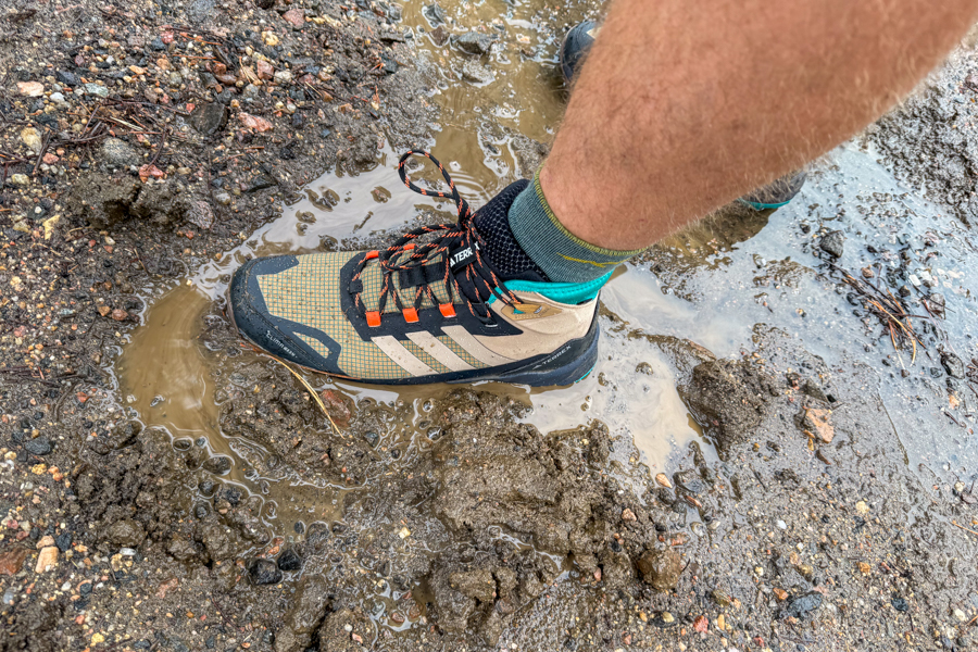 A close-up of a hiker’s foot wearing the Adidas Terrex Skychaser AX5 Mid GORE-TEX Climawarm+ shoe stepping into a muddy puddle on a wet trail.