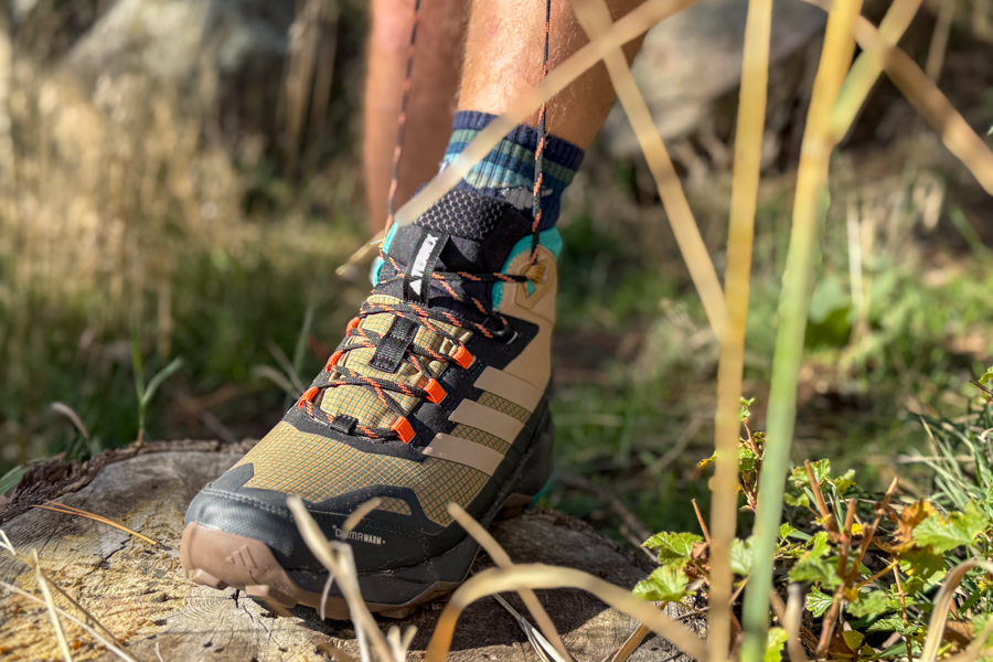 A hiker wearing the Adidas Terrex Skychaser AX5 Mid GORE-TEX Climawarm+ shoe steps on a tree stump in a sunlit grassy forest.