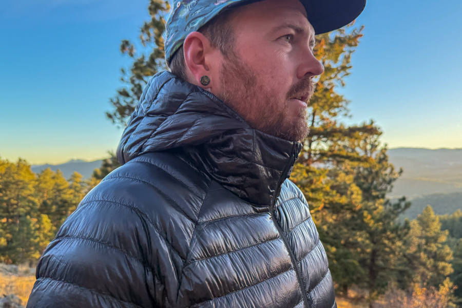 Profile view of the hiker wearing the Deploy Down 0.5 with the hood up, highlighting its snug fit and high collar against the sunset backdrop.