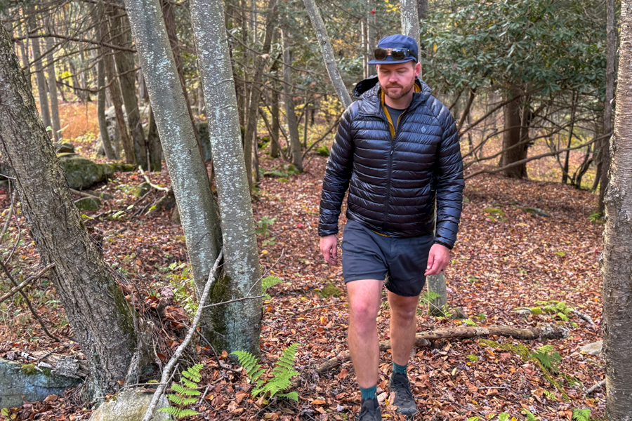 A hiker wearing the Black Diamond Deploy Down 0.5 Full Zip Hoody walks through a wooded trail covered in fall leaves, surrounded by trees and soft morning light.