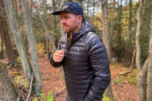 A man wearing the Black Diamond Deploy Down 0.5 Full Zip Hoody zips up the front of the jacket while hiking through a colorful fall forest.
