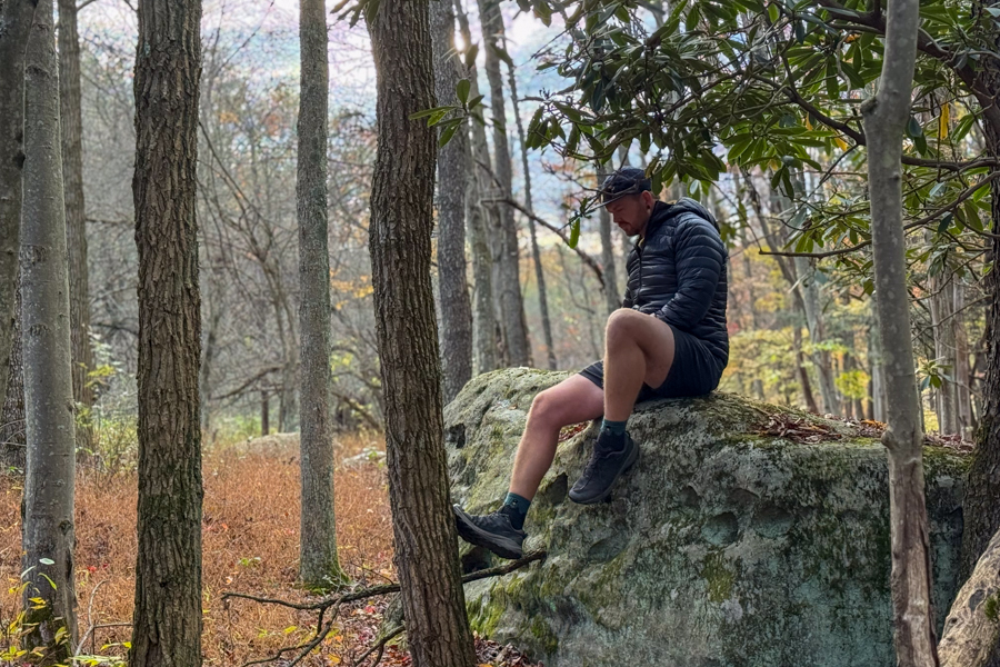 A hiker sits on a moss-covered boulder in the woods, resting in the ultralight puffy jacket with trees and golden undergrowth around him.