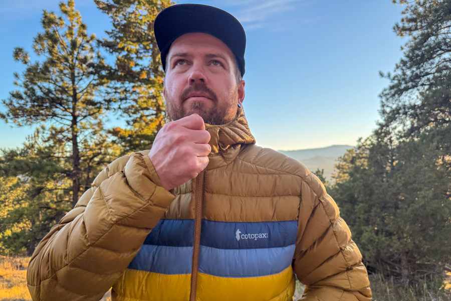 Close-up of a man zipping the Cotopaxi Fuego jacket while standing in golden light among pine trees.