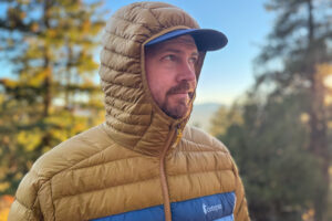 A man wearing the Cotopaxi Fuego jacket with the hood up, looking off toward the horizon in early morning light.
