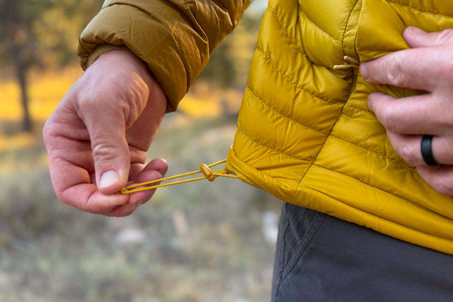 Close-up of a hand adjusting the elastic hem drawcord on the Cotopaxi Fuego Hooded Down Jacket to tighten the fit.