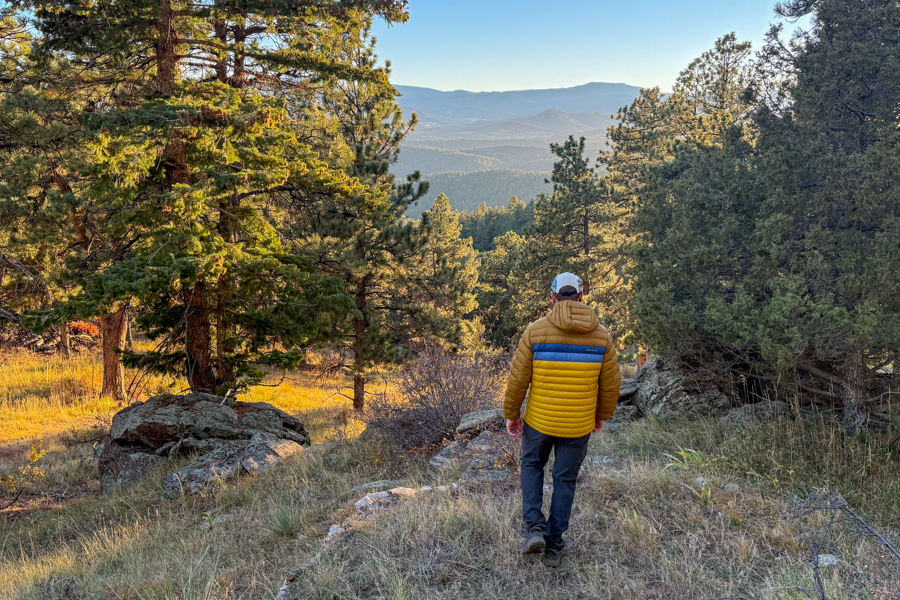 A man walking away through a forested overlook wearing the Cotopaxi Fuego jacket, with warm light illuminating the trees around him.