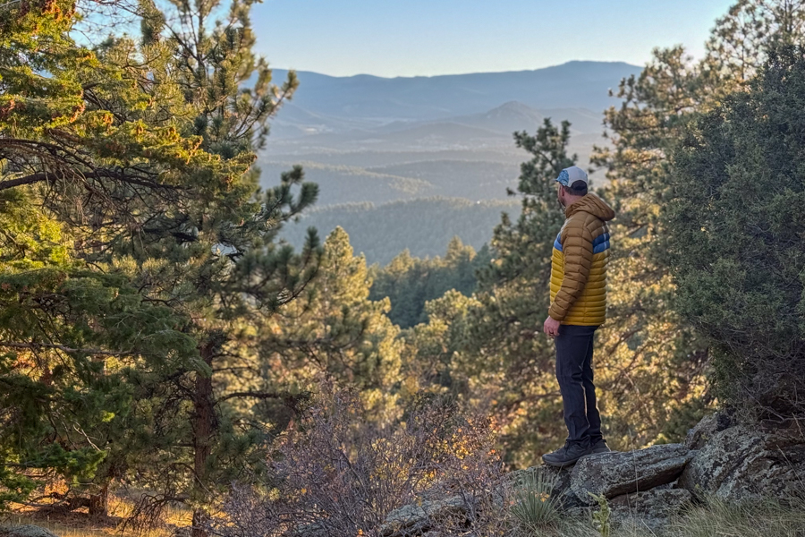 A man standing on a rocky overlook, gazing at mountain ridges under soft morning light while wearing the Cotopaxi Fuego jacket.