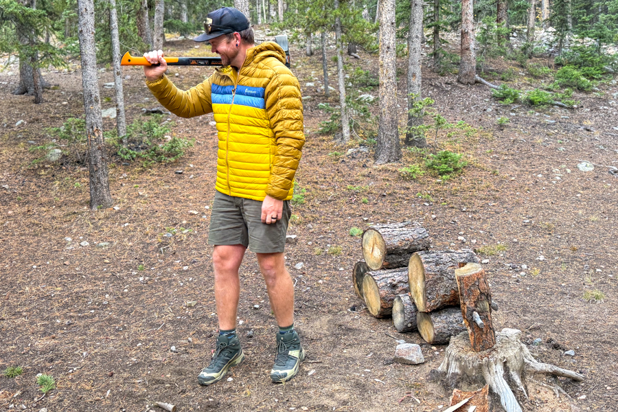 A camper wearing the Cotopaxi Fuego Hooded Down Jacket prepares to chop firewood in a pine forest campsite.