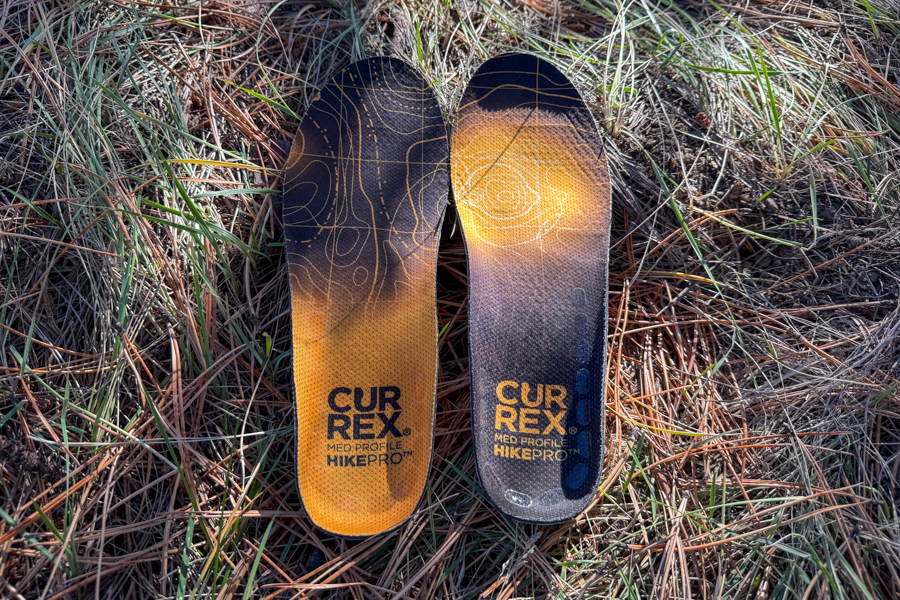 Top view of Currex HikePro insoles lying on dry grass with bold orange and black topo pattern graphics.