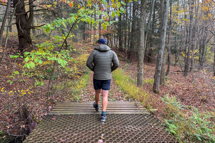 Rear view of a person walking through a wooded trail wearing the Simond MT100 Down Jacket, showing its length and quilted construction in motion.
