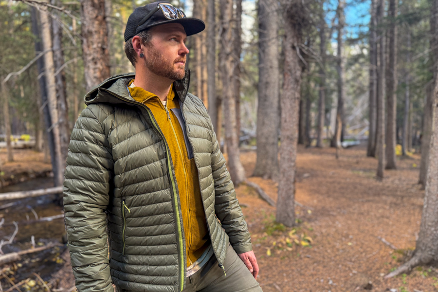 A person wearing the Simond MT100 Down Jacket and mustard midlayer stands near a forest stream, illustrating the fit and warmth for cool weather hikes.