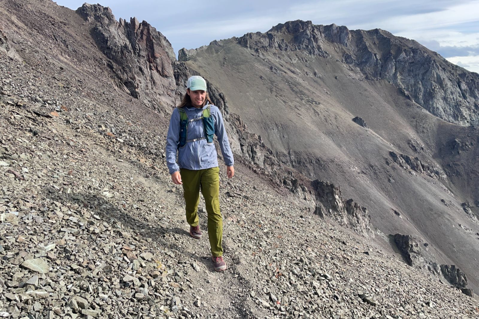 A man walks along a narrow trail on a rocky ridge wearing green pants.