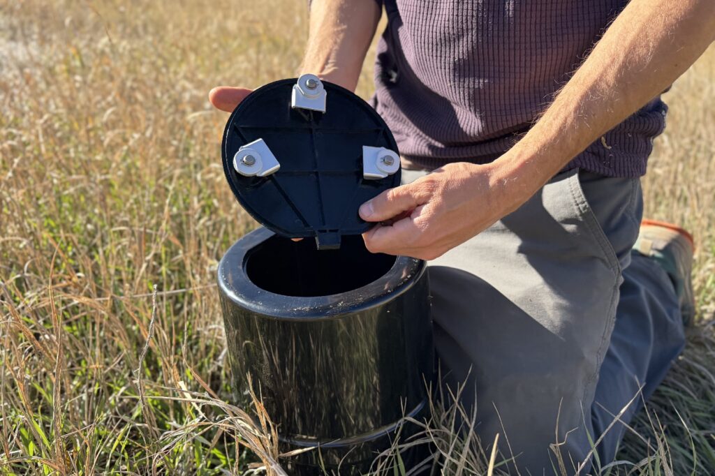 a hiker shows the underside of a bear canister lide while kneeling in the grass