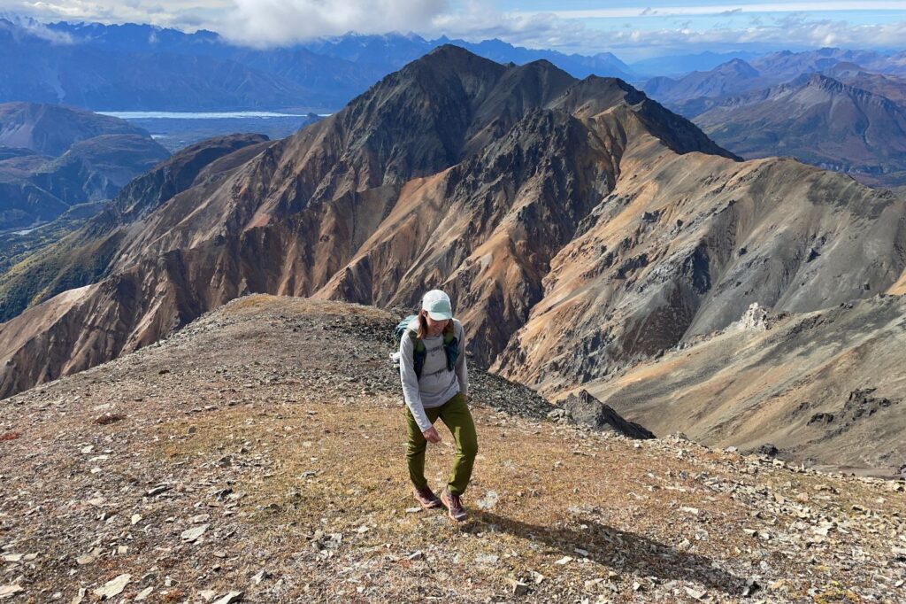 A man walks up a tundra slope in the alpine with orange colored scree ridges in the background.
