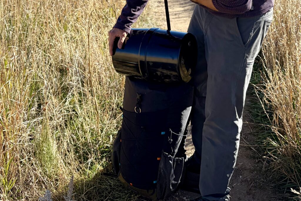 a hiker holds a black bear canister on top of a backpack while stretching a strap over top