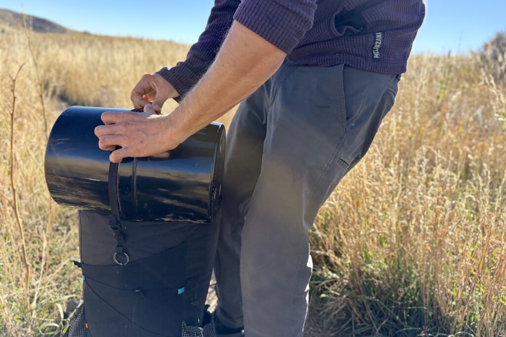 a hiker straps a bear canister to the top of a backpack