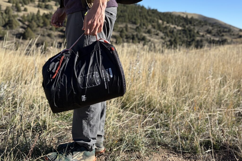 a waist down view of a hiker standing and holding a bear canister by its carrying case