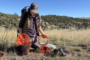 a hiker kneels on the ground while holding two separate parts of an orange bear canister
