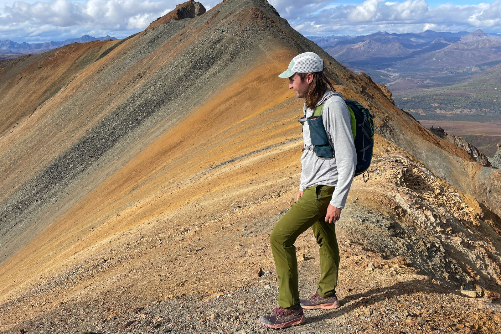 A man stands on a ridgeline made up of orange rock while wearing green pants.