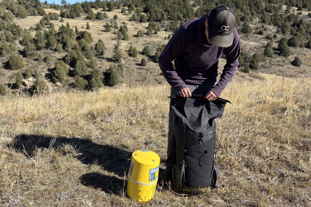 a hiker rolls up his backpack next to a yellow bear canister