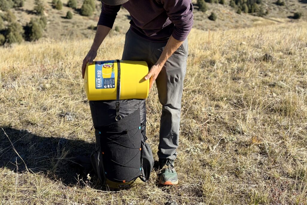 a hiker holds a backpack on the ground with a bear canister on top